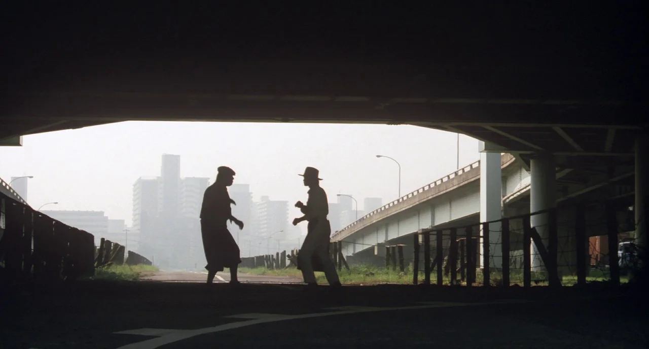Tampopo (1985) movie still: wide — An urban industrial space beneath a concrete road overpass looking out toward city buildings. Two men stand…