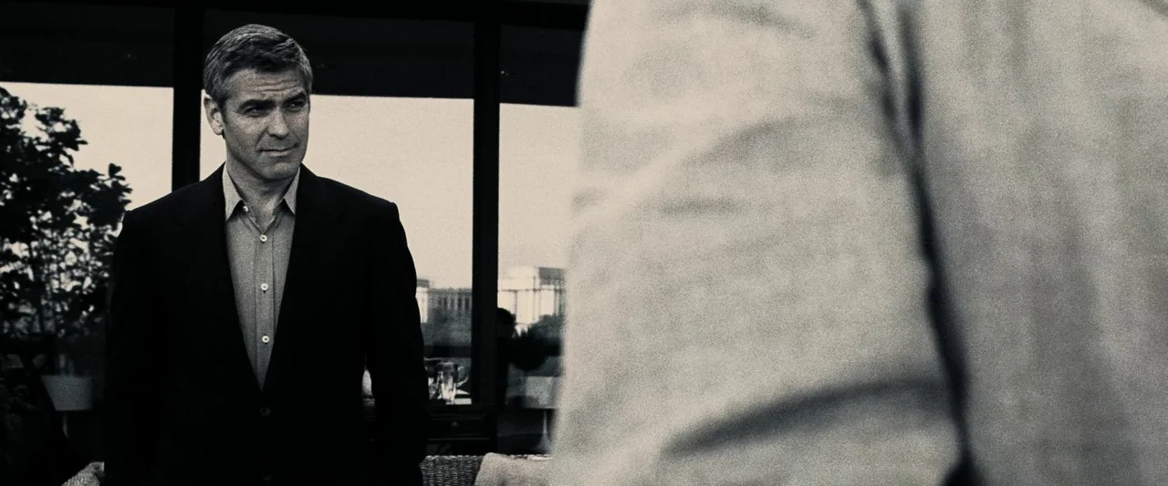 Ocean’s Twelve (2004) movie still: medium — A modern room features a large window backdrop with blurred architecture visible outside. A man in a…
