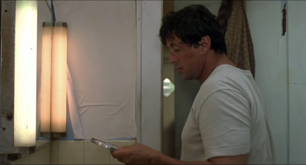 Cop Land (1997) movie still: medium — A small bathroom with cream-colored tiled walls and a vertical light fixture. A middle-aged man in a white…
