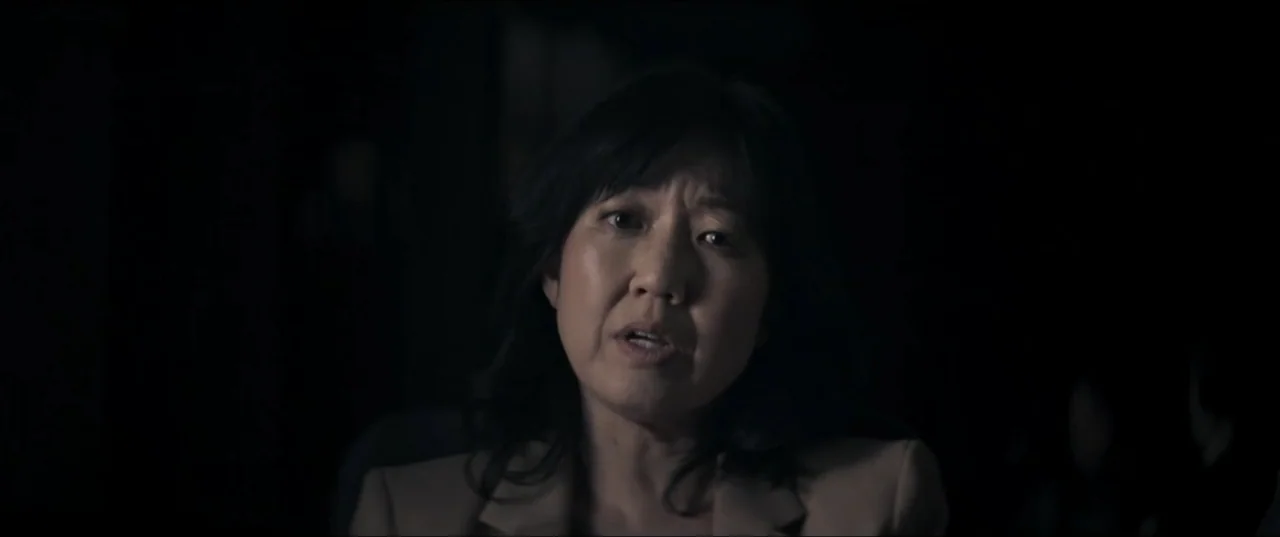 Offseason (2022) movie still: closeup — A person sits in a dimly lit space with a dark, indistinct background. A woman in a beige blazer speaks…