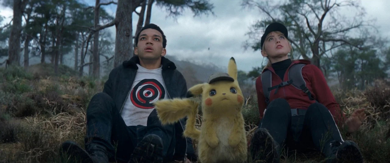 Pokémon Detective Pikachu movie still: medium — A sparsely wooded clearing with tall pine trees under an overcast sky. A young man, a young woman,…