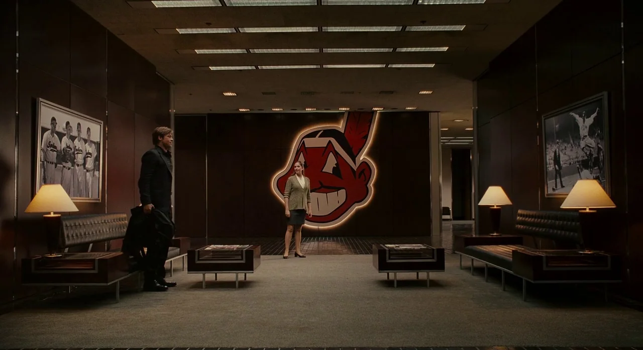 Moneyball (2011) movie still: wide — A spacious, dark-paneled lobby features low leather furniture and a large red mascot emblem. A man stands near…