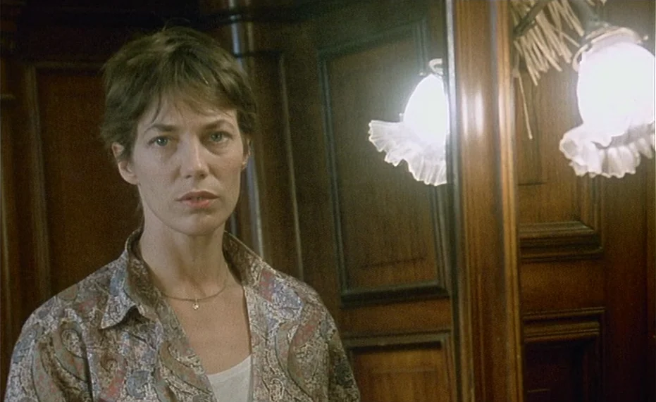 Jane B. par Agnès V. (1988) movie still: medium — A woman stands in a wood-paneled room beside a wall mirror with attached lamps. She gazes directly…