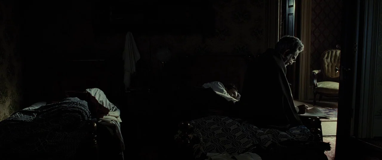 Lincoln (2012) movie still: wide — A dark, dimly lit domestic space features beds draped with heavy patterned blankets. An elderly man sits on the…