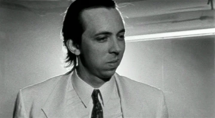 Roadkill (1989) movie still: closeup — A man stands in front of a plain, brightly lit indoor wall. The figure occupies the center of the frame…
