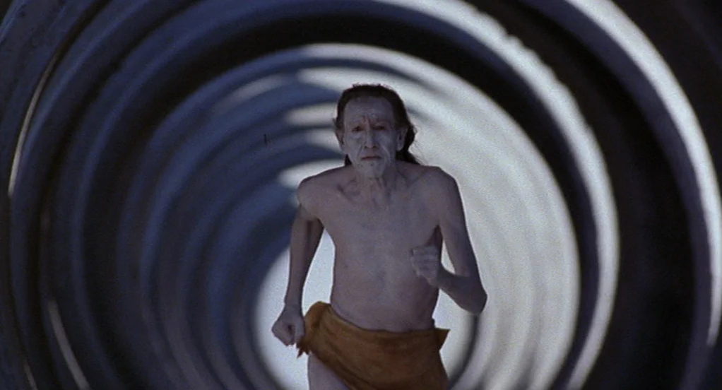 The Indian Runner (1991) movie still: medium — A thin, pale-skinned man runs through the interior of a large corrugated metal pipe. The circular…