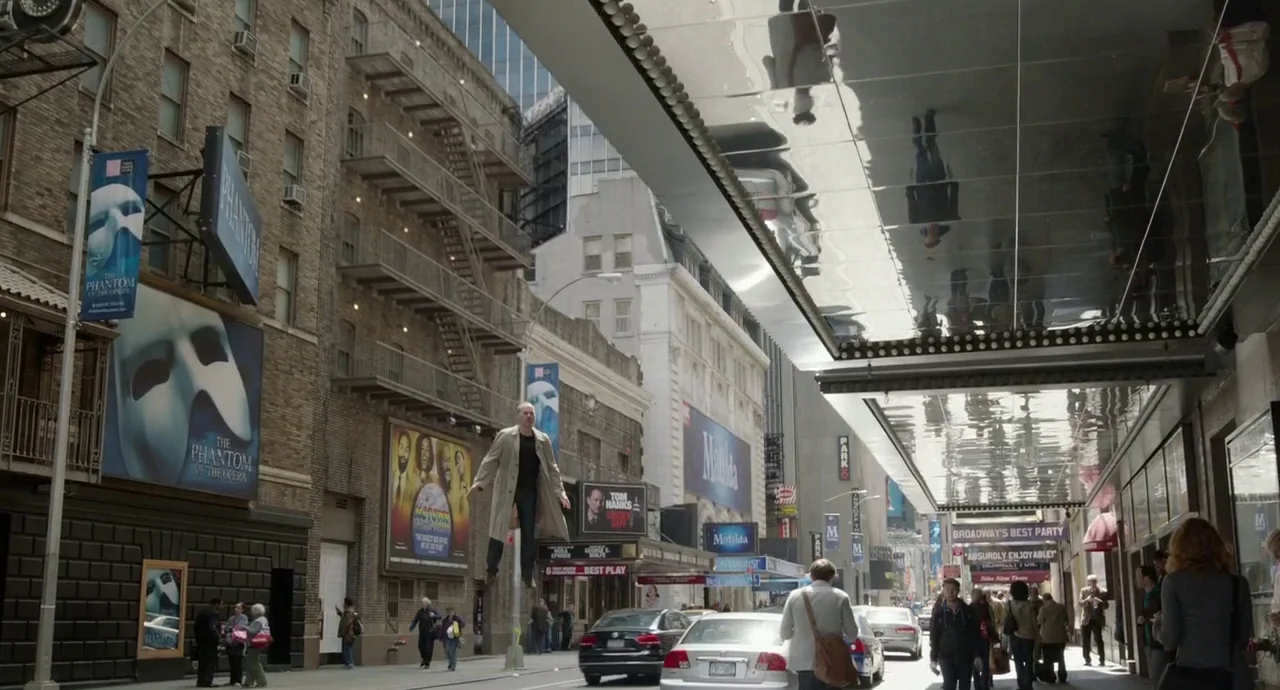 Birdman (2014) movie still: wide — A bustling New York City street lined with Broadway theater posters and billboards. A man in a trench coat…