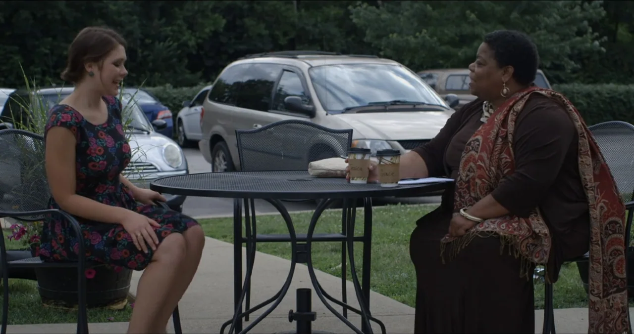 Faith, Love & Chocolate (2018) movie still: medium — A metal patio table sits on a concrete sidewalk in front of a parking lot. Two women sit across…