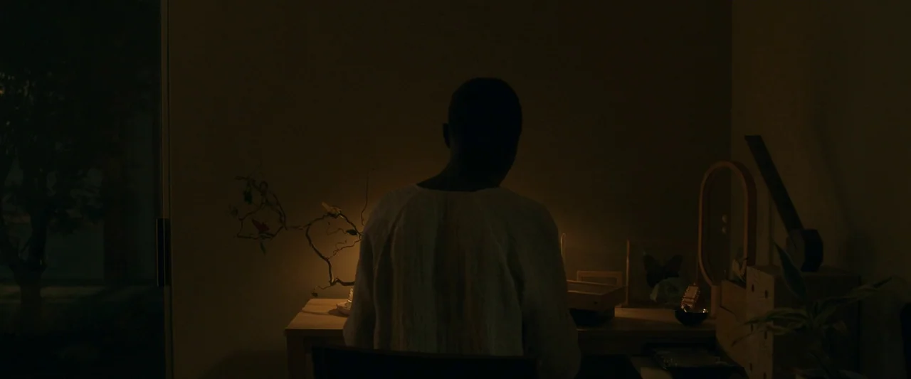 After Yang (2021) movie still: medium — A dark, dimly lit room contains a person seated at a simple wooden table. The desk is sparsely decorated…