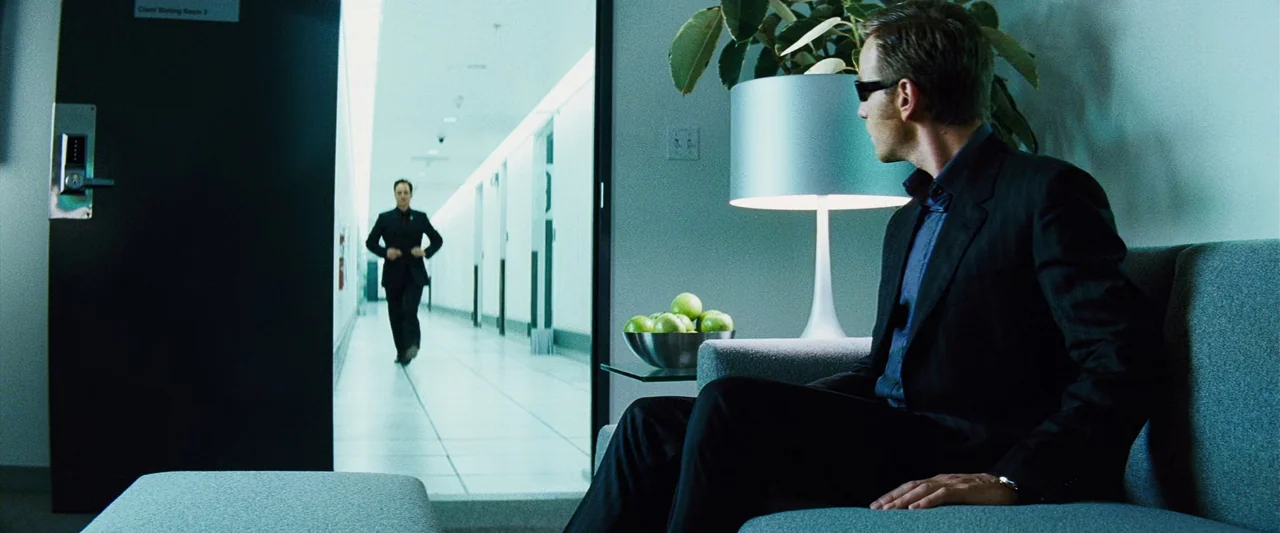 The Island (2005) movie still: medium — A modern, starkly lit waiting room with white tile floors and white walls. A man in a dark suit sits in the…