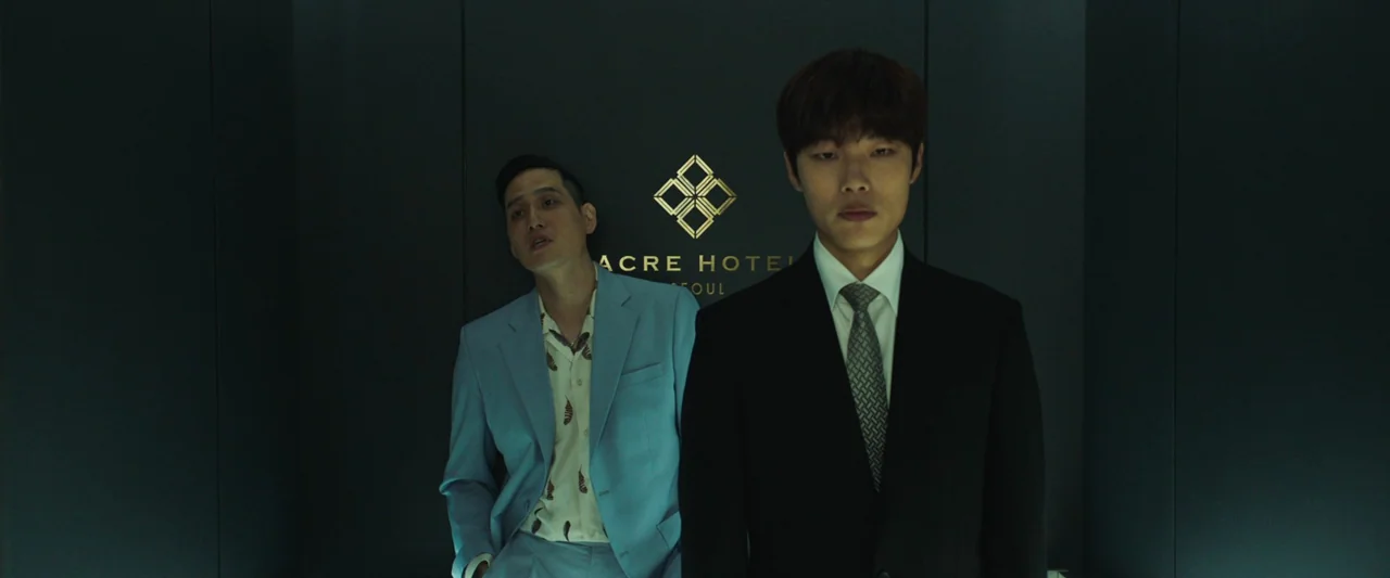 Believer (2018) movie still: medium — A dark hotel reception wall displays a gold geometric logo above the text Acre Hotel. Two men stand in front…
