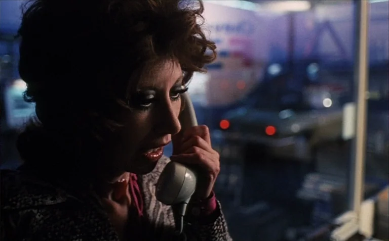 Business is Business (1971) movie still: closeup — A dimly lit phone booth features blue-tinted windows reflecting distant blurred red street…