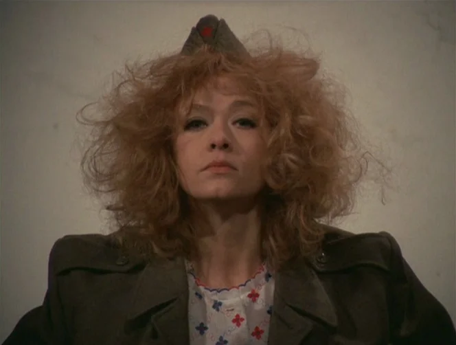 WR: Mysteries of the Organism (1971) movie still: closeup — A woman stands against a featureless, light-colored wall in a sparsely lit space. She…