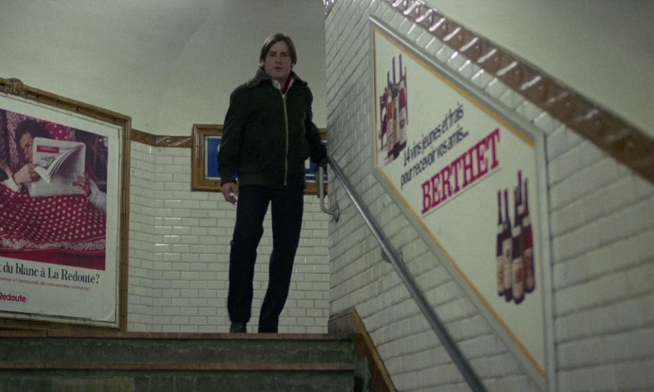 The Margin (1976) movie still: fullbody — A subterranean subway staircase features white subway tiles and large vintage French advertisements. A man…