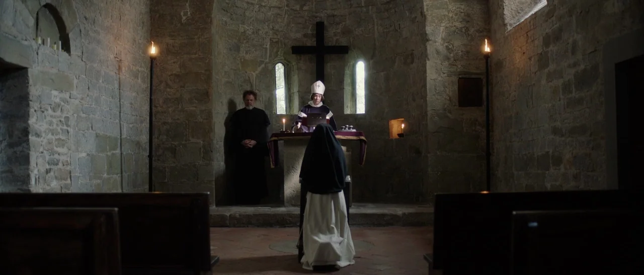 The Little Hours (2017) movie still: wide — A rustic stone interior with thick walls and a simple altar under a cross. A nun kneels in prayer while…