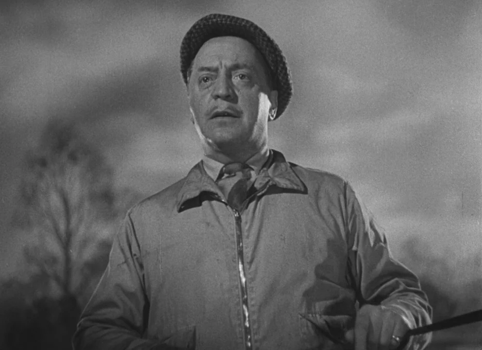 Dead of Night (1945) movie still: medium — An open field under a cloudy sky serves as the backdrop for a man. The man in a flat cap stands against…