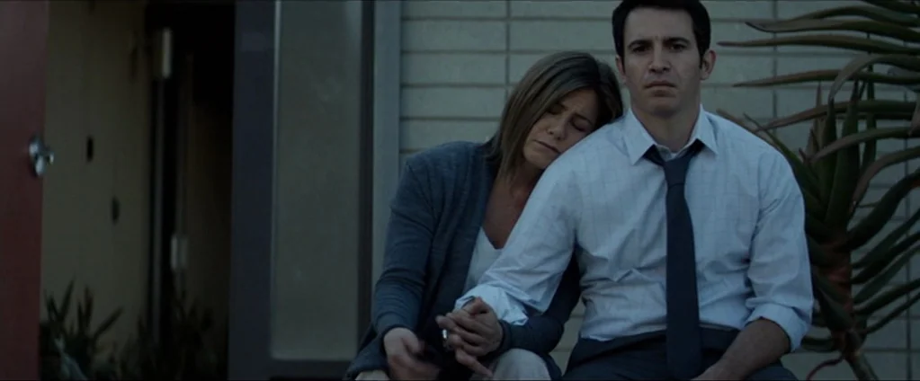 Cake (2014) movie still: medium — A residential exterior at dusk with a brick facade and a decorative plant. A woman rests her head on a man's…