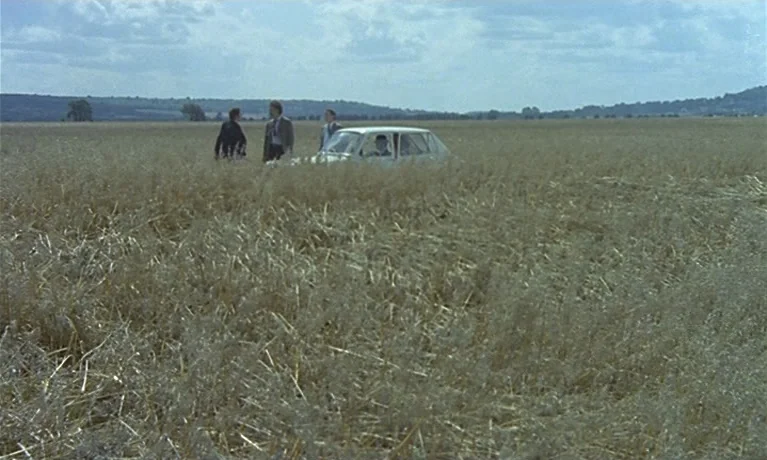 Nuits Rouges (1974) movie still: wide — A vast, dry grassy field extends to the horizon under a cloudy blue sky. Three men stand beside a white…