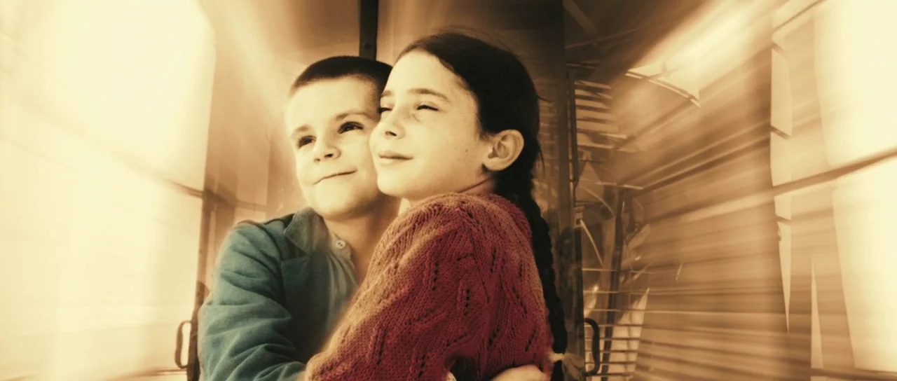 A Very Long Engagement (2004) movie still: medium — A pair of children stand inside a train carriage with windows revealing blurred motion. The boy…