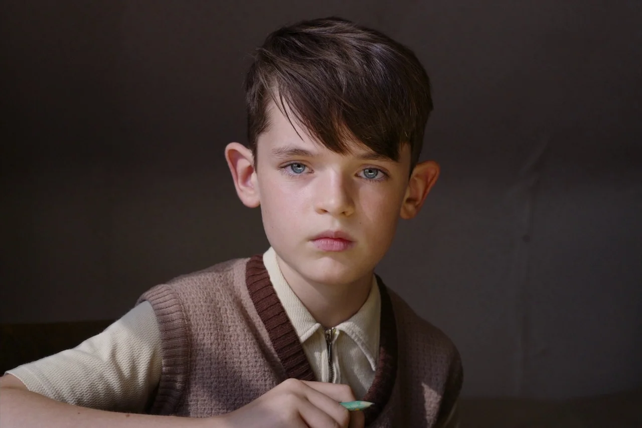 Anselm (2023) movie still: closeup — A young boy sits in a dimly lit, nondescript room. He holds a pencil while facing directly toward the camera…