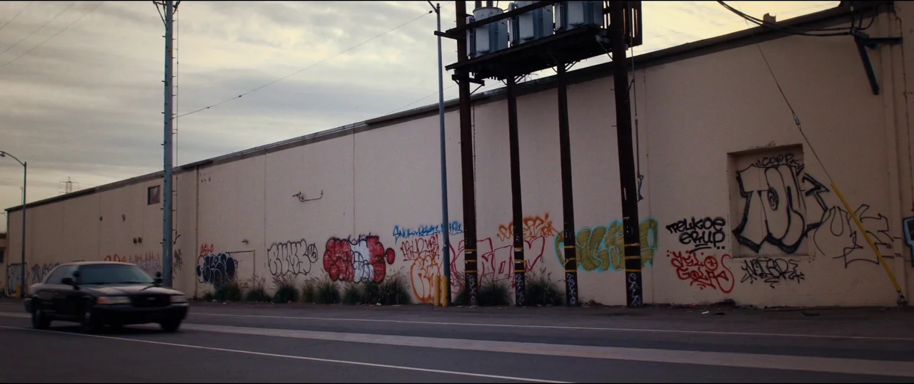 El Chicano (2018) movie still: wide — A long, cream-colored windowless building wall features extensive colorful graffiti markings along its base. A…