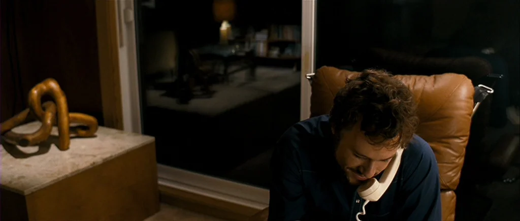I’m Not There (2007) movie still: medium — A modern living room interior at night with a large glass pane wall. A man sits in a leather chair while…