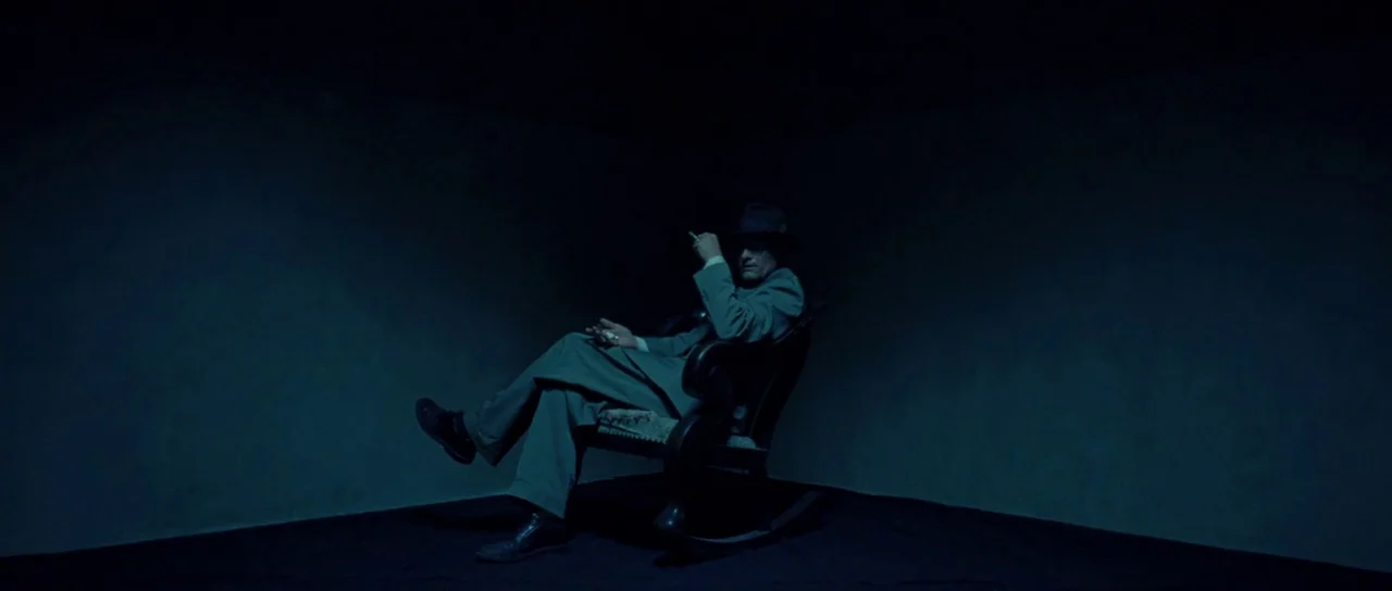 On The Road (2012) movie still: fullbody — A person sits in a simple wooden rocking chair inside a dark, empty corner. The minimalist space has deep…