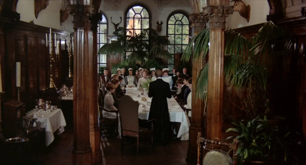 The Go-Between (1971) movie still: wide — A formal dining room features dark wood paneling and large arched windows with plants. A group of people…