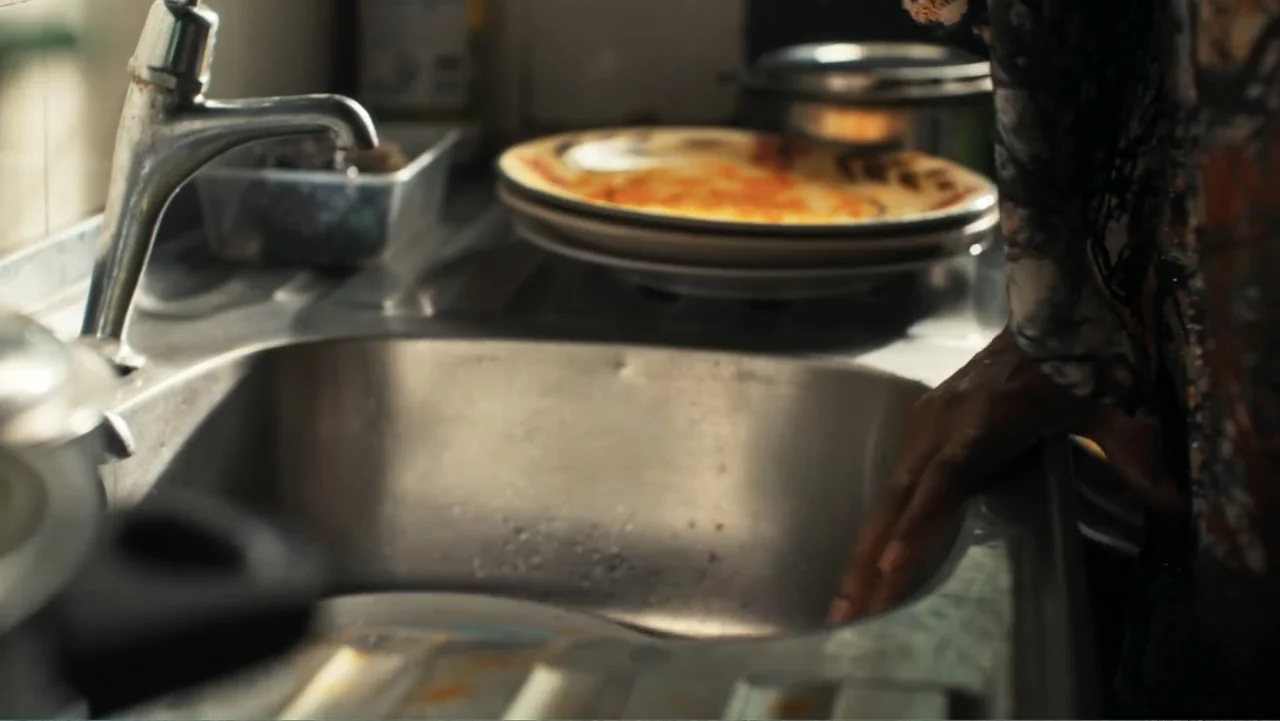 Gangs of Lagos (2023) movie still: medium — A cluttered kitchen sink area with a metallic faucet and a stack of plates. A person's hand rests on the…