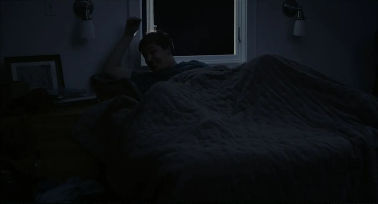 Freedom (2024) movie still: medium — A dimly lit bedroom features a bed with a dark textured quilt. A young man sits up in bed reaching toward a…