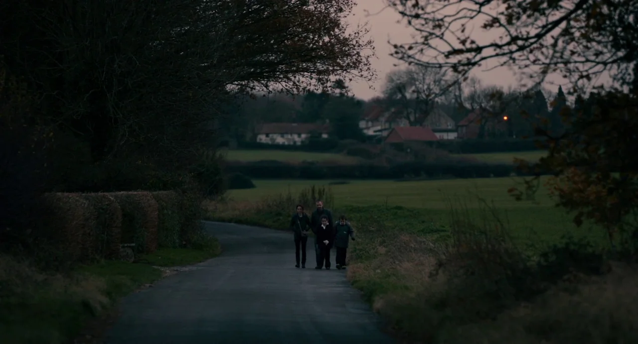 Everyday (2012) movie still: wide — A quiet rural road flanked by hedges and fields leads toward distant houses. Three people walk side-by-side…