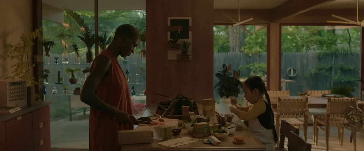 After Yang (2021) movie still: wide — A modern kitchen features wood paneling and extensive glass walls looking out to greenery. A woman slices…