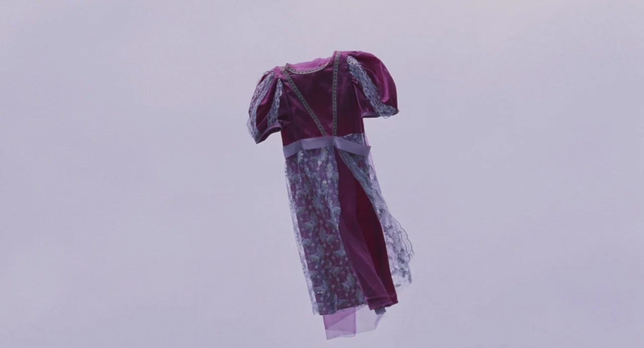 Ma Vie En Rose (1997) movie still: closeup — A magenta child's dress with lace trim floats against an overcast, hazy sky. The garment hangs in a…