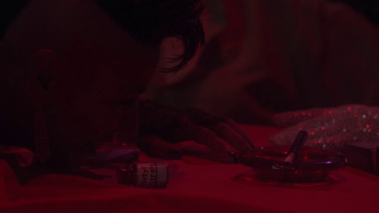 Repo Man (1984) movie still: closeup — A dark tabletop covered in a red cloth holds a small bottle and ashtray. A person with a dark spiked haircut…
