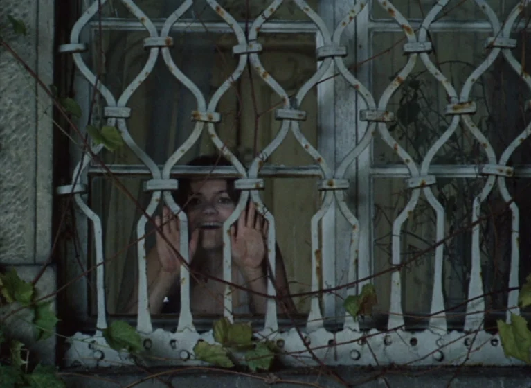 Fruit of Paradise (1970) movie still: medium — A woman looks through a weathered, white metal security grate covering a window. Vining green plants…