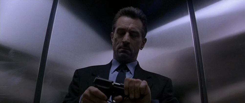 Heat (1995) movie still: medium — A man stands inside a stainless steel elevator with clean, reflective panels. He looks down at a black pistol…