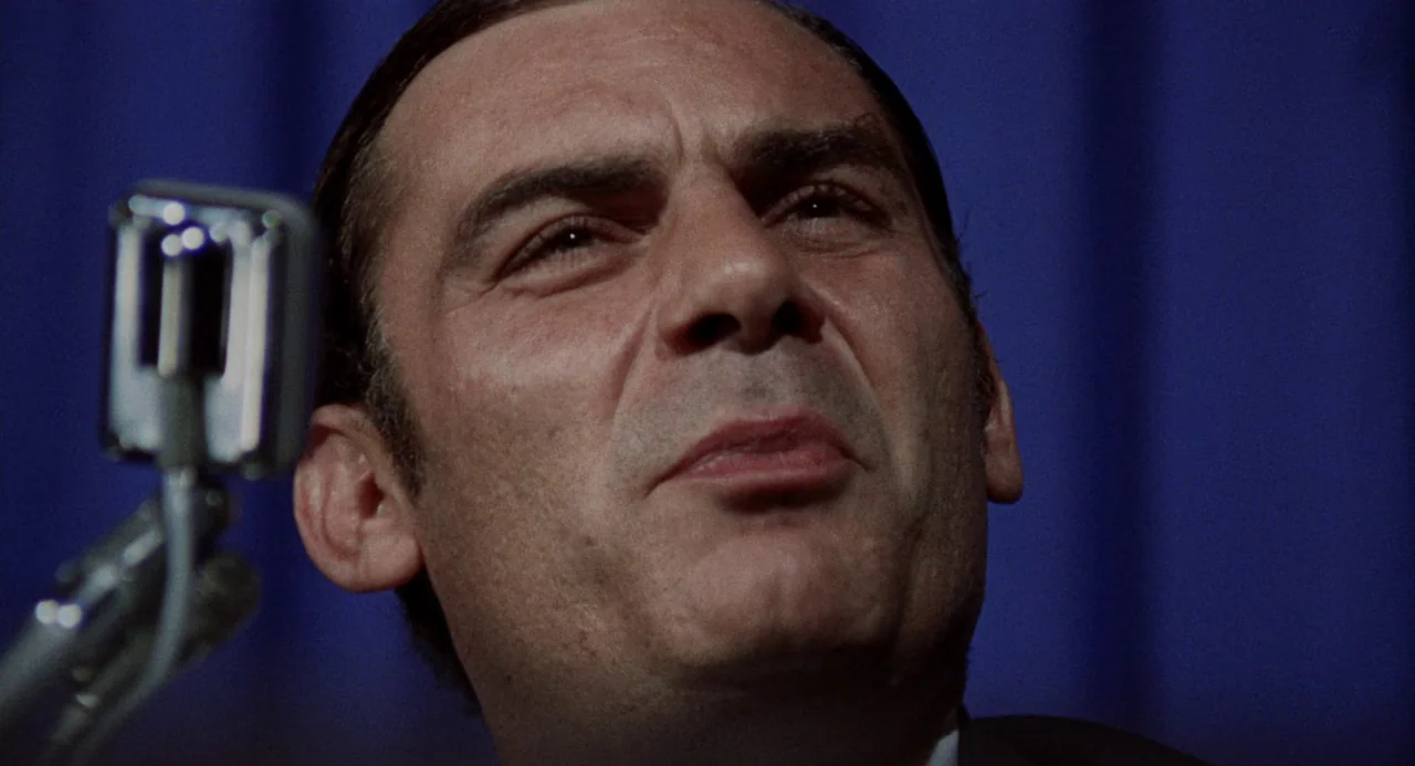 Investigation of a Citizen Above Suspicion (1970) movie still: closeup — A man speaks at a press conference against a dark blue curtain backdrop. He…