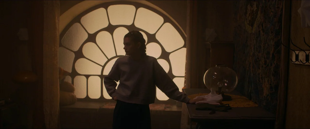 Birds of Paradise (2021) movie still: medium — A dimly lit room features a large arched window with a segmented decorative pane. A young woman…