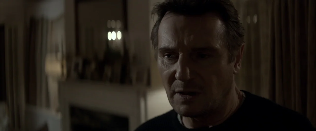 A Walk Among the Tombstones (2014) movie still: closeup — A dimly lit room features a blurred fireplace area in the background. A man stands in the…