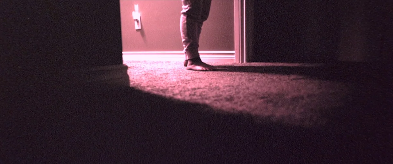Skinamarink (2022) movie still: wide — A dark, carpeted floor leads into a narrow hallway illuminated by a small plug-in light. Bare feet stand…