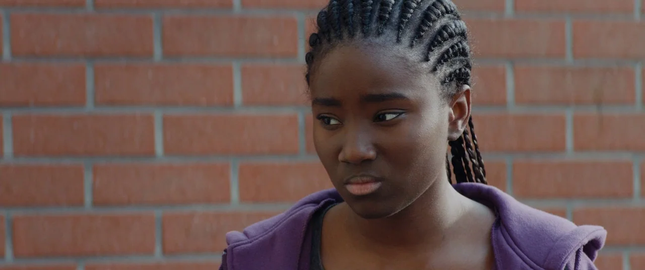 Girlhood (2014) movie still: closeup — A young woman stands in front of a flat, orange-red brick wall exterior. She looks slightly to the side with…