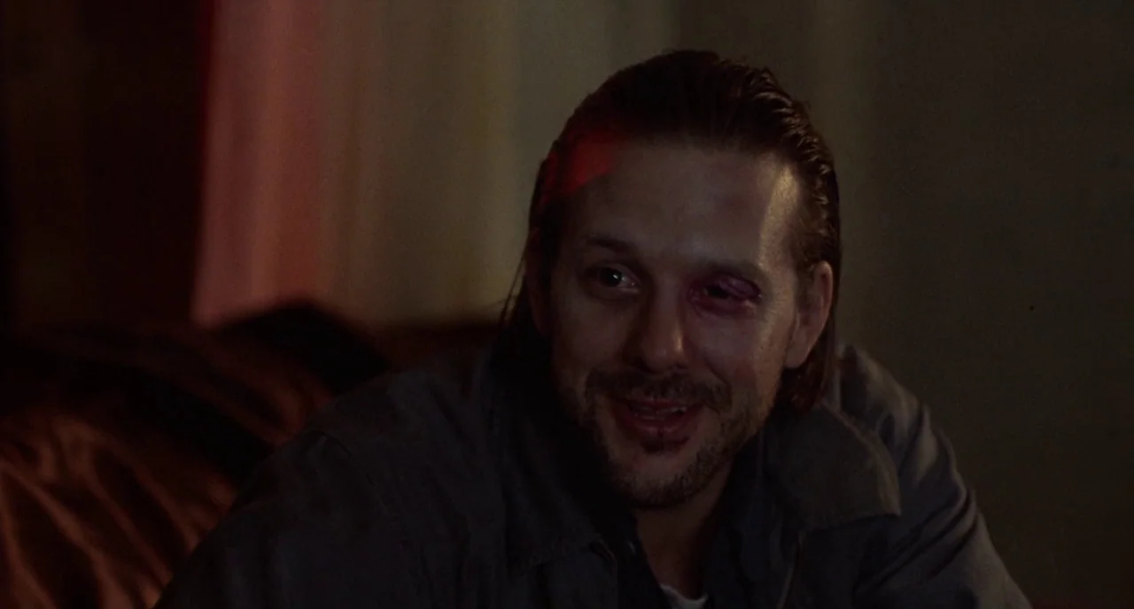 Barfly (1987) movie still: closeup — A dimly lit indoor space with indistinct shadows forming the background. A man with a bruised eye and slicked…