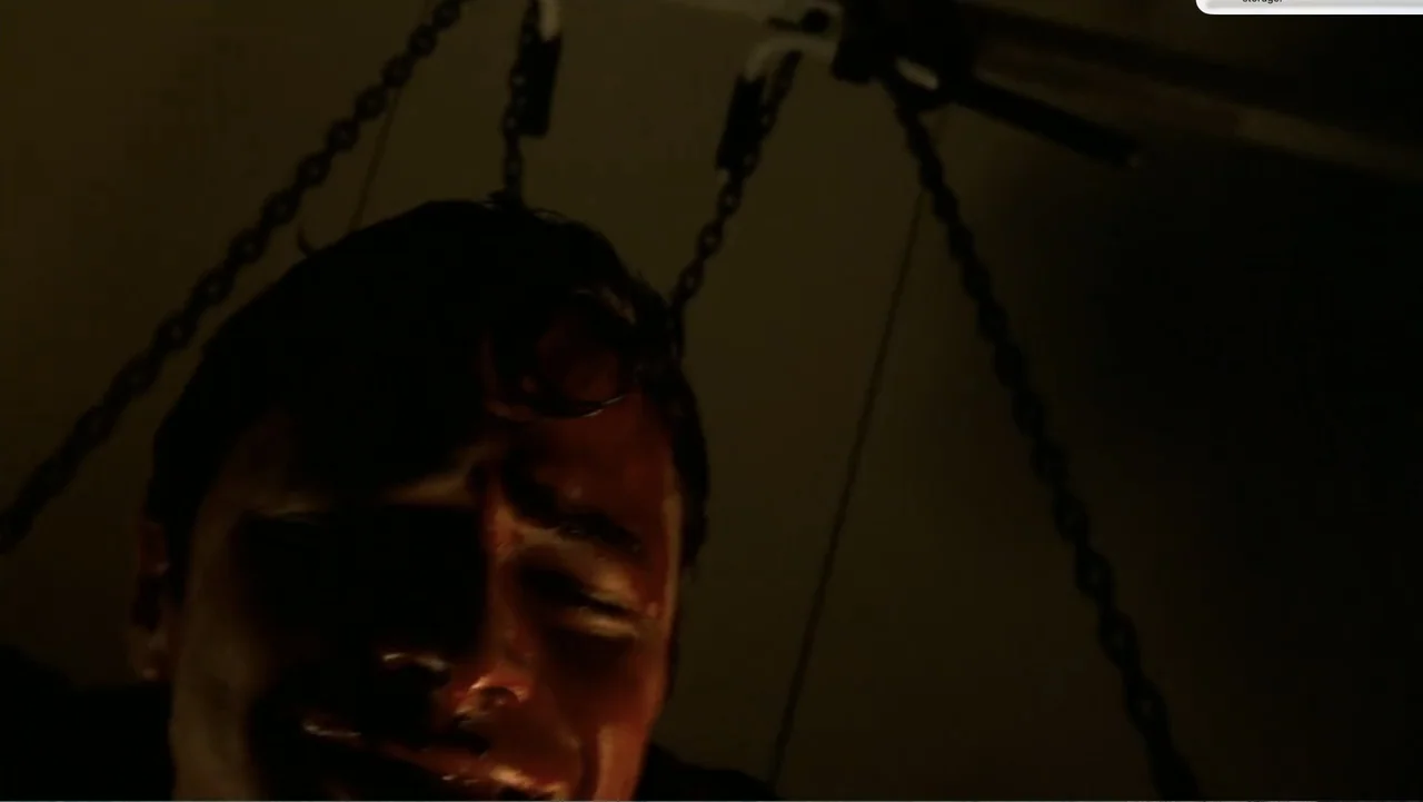 Chain Letter (2010) movie still: closeup — A dark indoor space features metal chains hanging prominently in the background. A man stands in the…