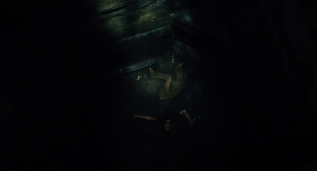 Submergence movie still: wide — A cramped, dark underground chamber with rough, uneven walls and a dirt floor. A single person lies on the ground,…