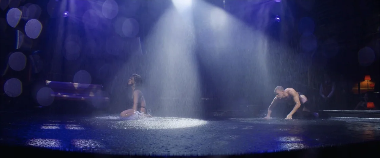 Magic Mike's Last Dance (2023) movie still: wide — A dark stage interior features an overhead spotlight and artificial rain falling down. A man and…