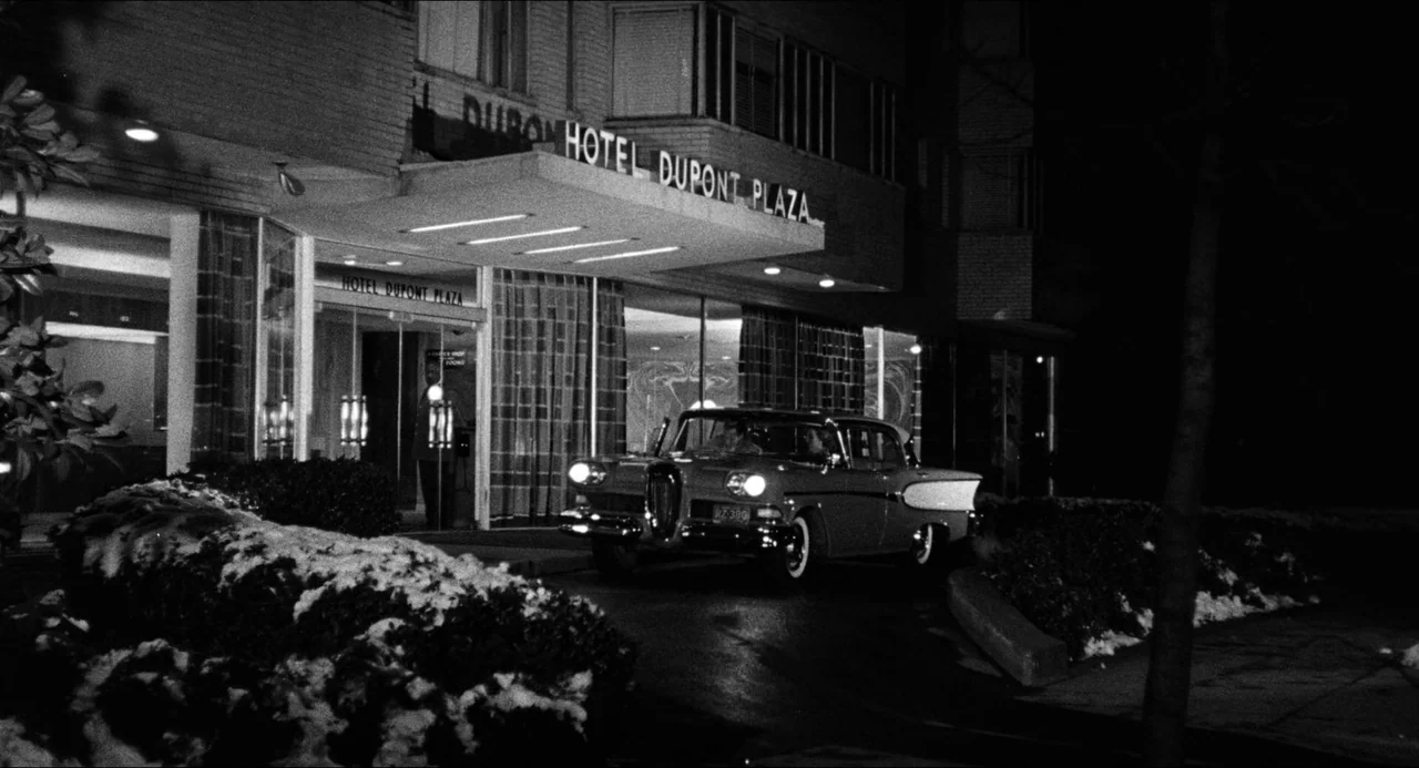 The Fearmakers (1958) movie still: wide — The exterior of a hotel building with a large illuminated sign at night. A mid-century car is parked under…