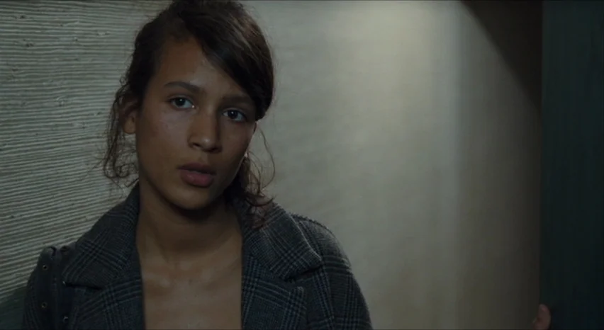 35 Shots of Rum (2008) movie still: closeup — A dimly lit indoor hallway with a textured, pale wall surface. A young woman stands facing the camera…