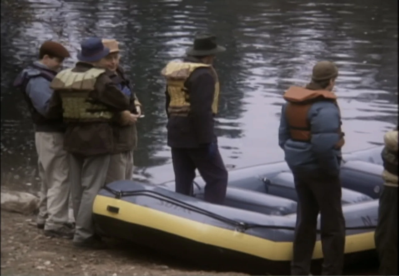 White Mile (1994) movie still: fullbody — A group of men stand on a pebbled bank next to a large rubber raft. They wear hats and life vests while…