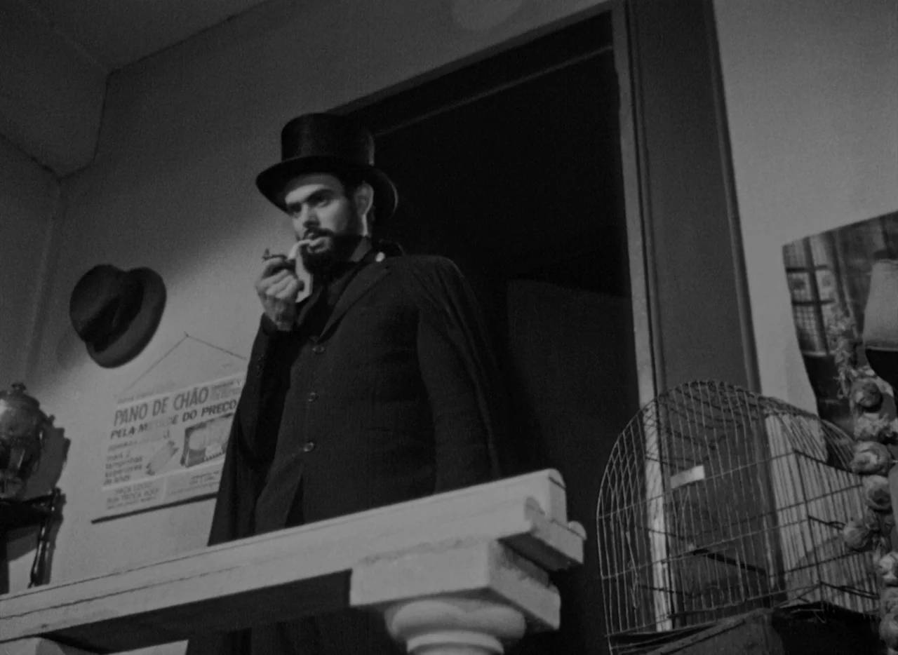 At Midnight I’ll Take Your Soul (1964) movie still: medium — A man in a top hat stands in a dim room with a birdcage nearby. He holds a pipe to his…