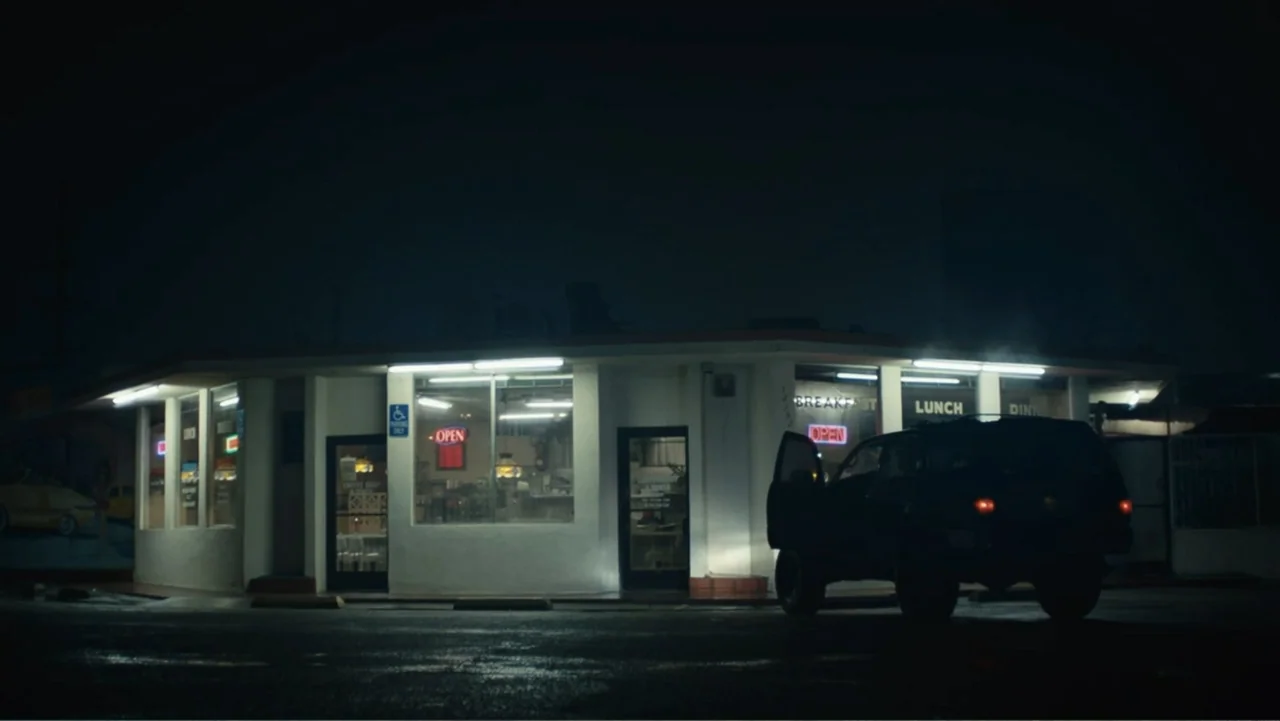 The Cleaner (2021) movie still: wide — A quiet roadside diner at night features bright fluorescent lights reflecting on wet pavement. A dark SUV…